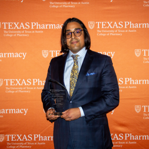 Mike Sandoval holds is award and smiles to camera. He has long hair and glasses and is wearing a dark navy suit and tie. Mike stands in front of an orange, Texas Pharmacy-branded wall.