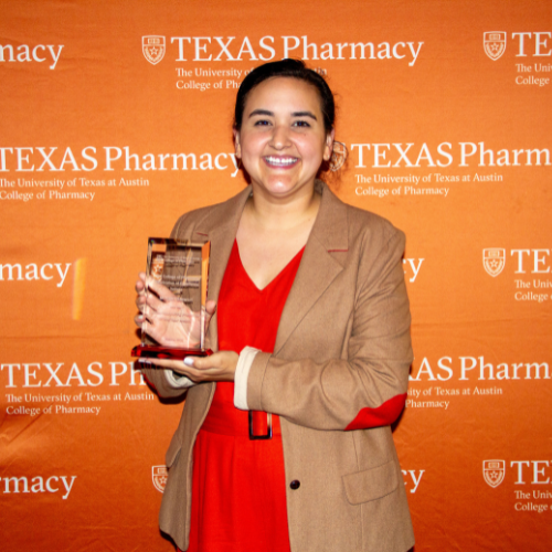 Dr. Stewart holding an award. She is wearing a red dress with a camel-colored jacket over top. She stands in front of an orange, Texas Pharmacy-branded wall.