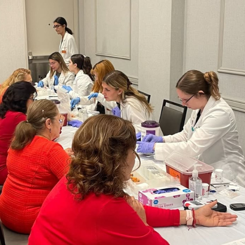 A group of Project Collaborate student pharmacists working at a screening event. The Pharm students wear white coats and gloves while the women being screened are in red tops.