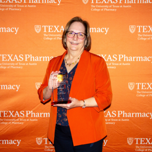 A photo of Professor Sharon Rush holding an award. She wears an orange jacket and stands infront of an orange Texas Pharmacy-branded wall.