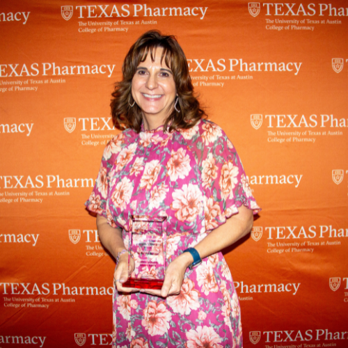 Dean Acosta holding an award. She is wearing a floral dress and is standing in front of an orange, Texas Pharmacy-branded wall.