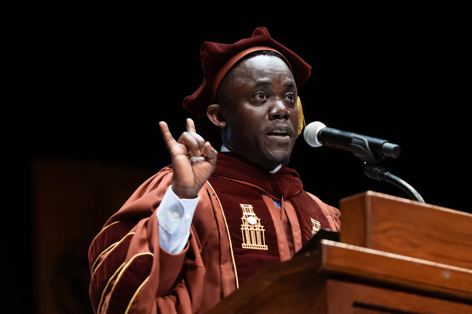 A man at a podium in regalia giving the Hook 'em Horns.