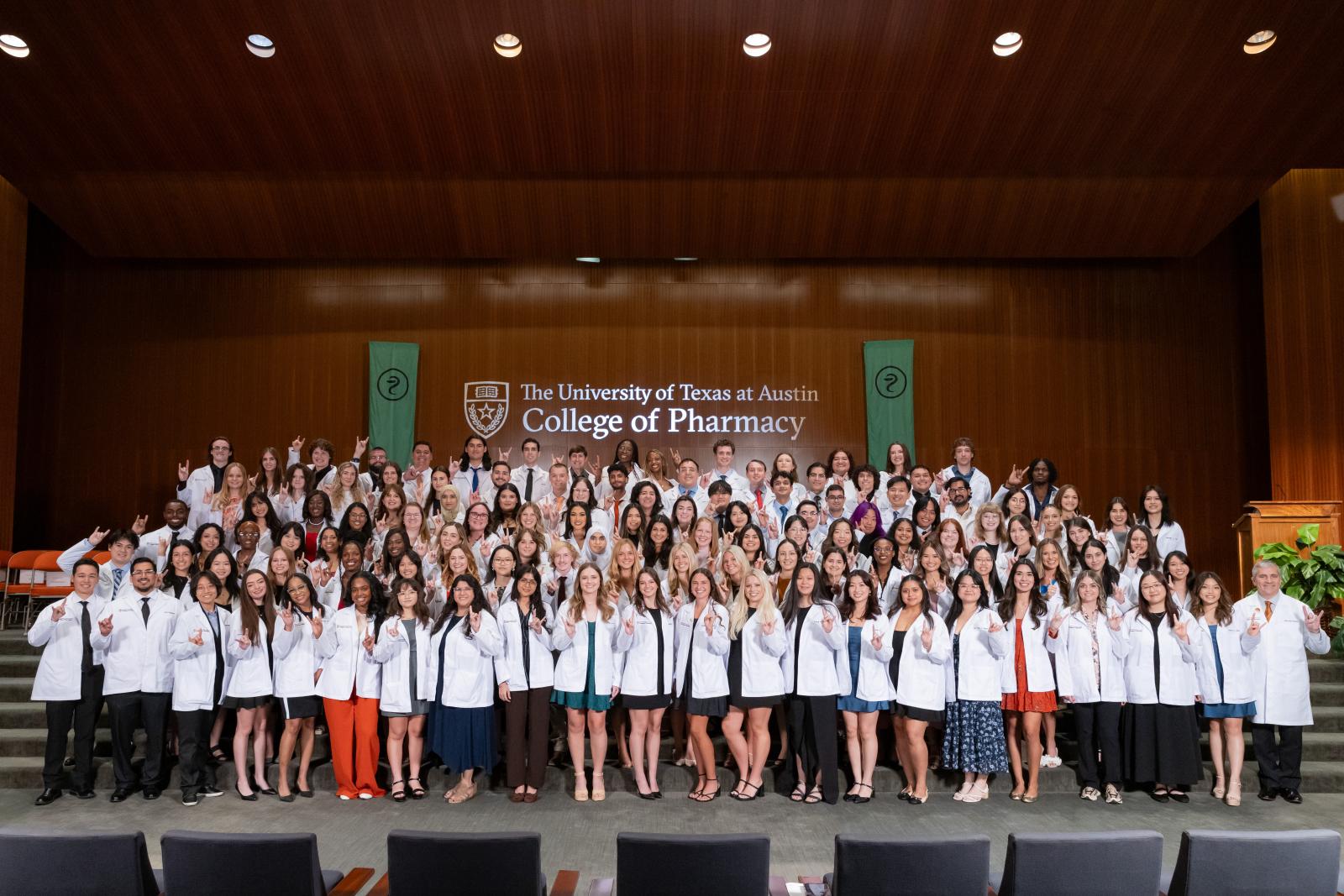 A class photo of the UT Austin College of Pharmacy Class of 2029. Students wearing their white coats stand on stage holding up the "hook 'em horns" hand gesture and smile to camera.