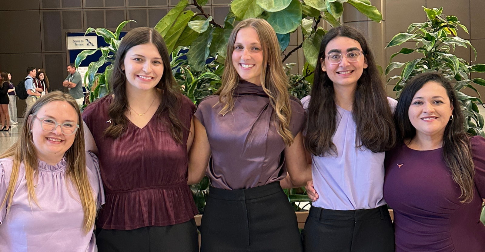 Five female student pharmacists pose together, smiling brightly and wearing purple business attire.