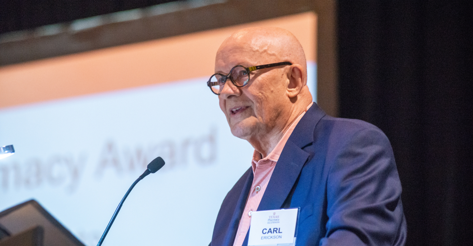 A man wearing a navy suit jacket and dark-rimmed glasses speaking into a microphone on stage.