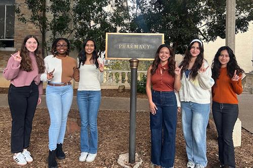 Six Pharm.D. students standing by the pharmacy building sign and giving the Hook 'Em Horns hand sign.