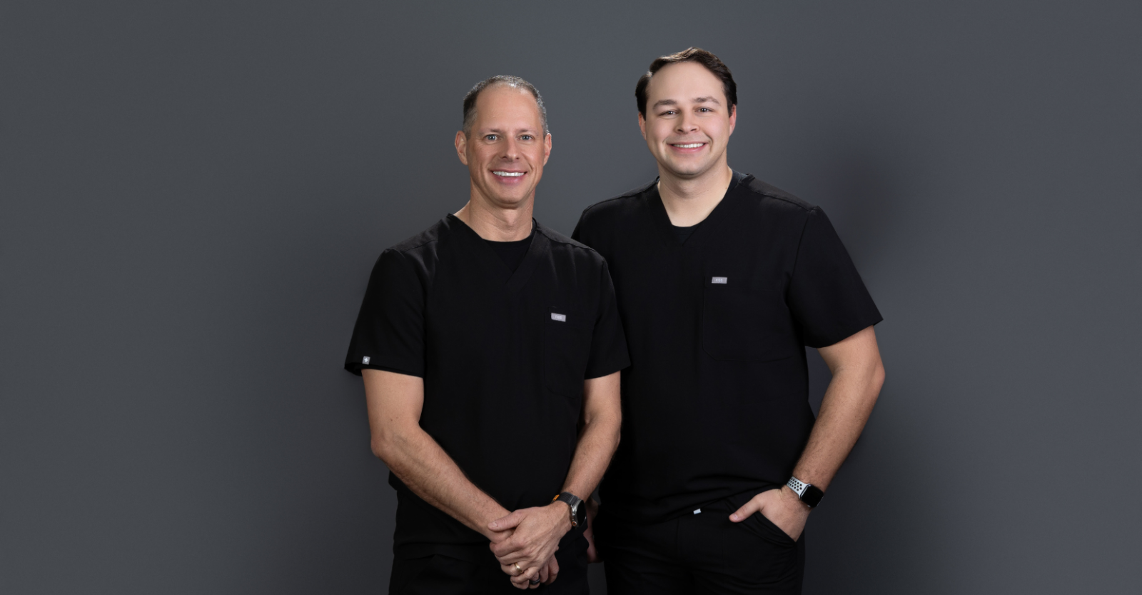 Father and son, Steve and Ryan Hoffart, stand together in black scrubs smiling to camera. They stand against a dark grey backdrop.