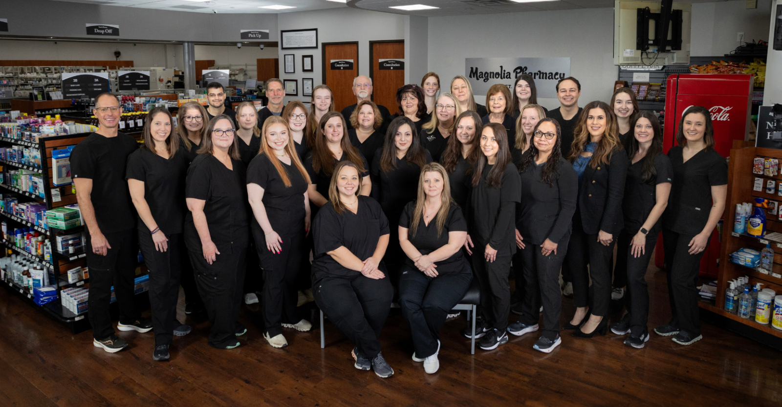A group photo of the staff from Magnolia Pharmacy. All are wearing black scrubs and smiling warmly to camera while standing inside the Pharmacy.