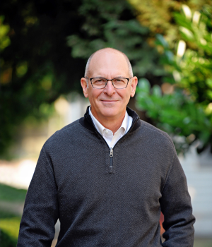 Headshot of Mike Lester. Mike stands outside and smiles to camera in a grey half-zip sweater and rectangular glasses.
