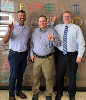 Three men, dressed professionally, pose together with the "Hook 'em Horns" hand gesture in front of a decorative wall.