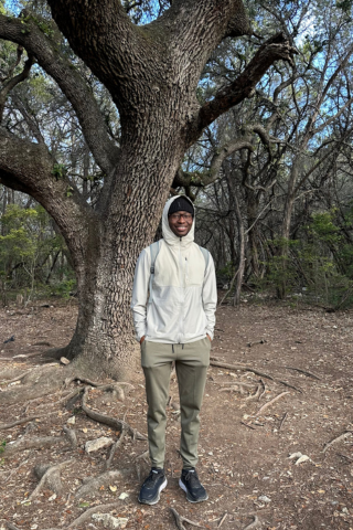 An adult male wearing outdoor walking gear poses on a trail in front of a large tree.