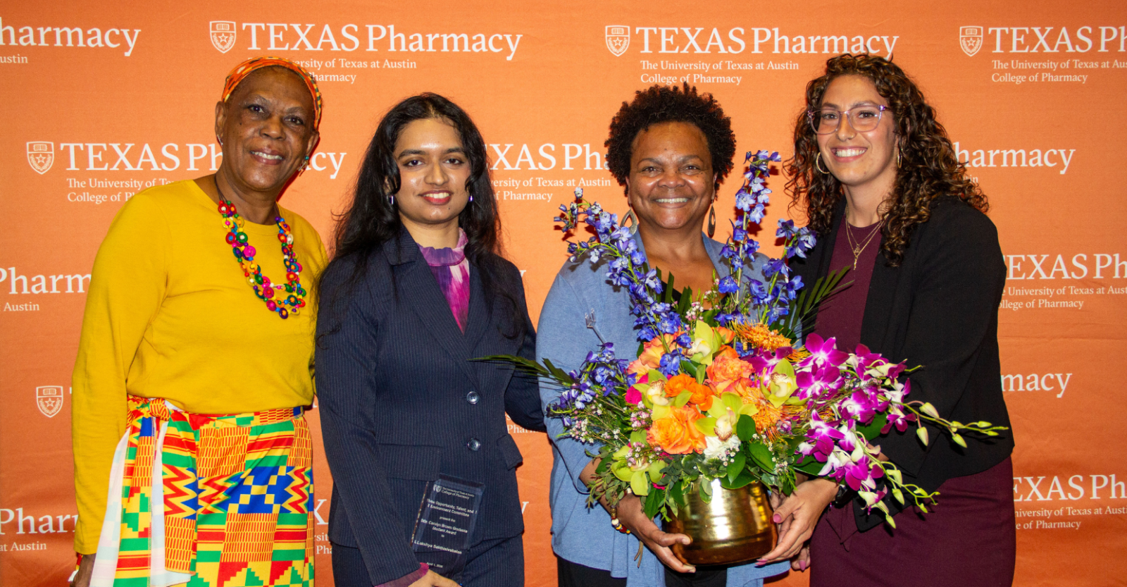 Four smartly dressed women pose together at UT Austin College of Pharmacy academic award ceremony. One central figure holds a bouquet of flowers, another an award, and all are smiling brightly to camera.