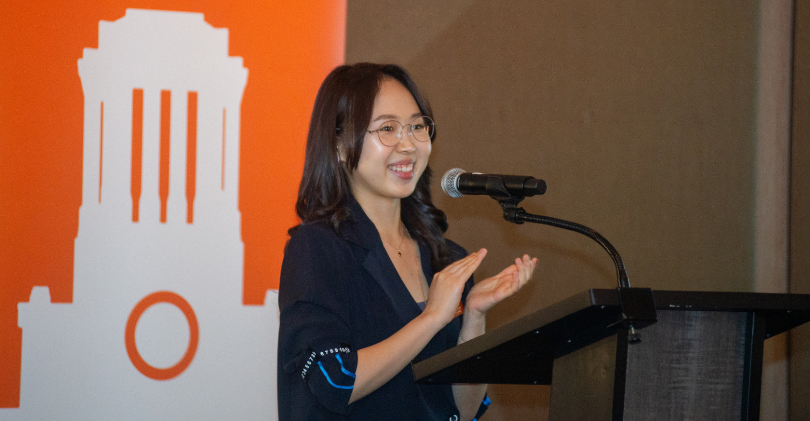 Pharmacy student, Doeun, on stage at a college event. Doeun is wearing a black blazer and glasses and is standing at a podium and microphone, hands up in applause.
