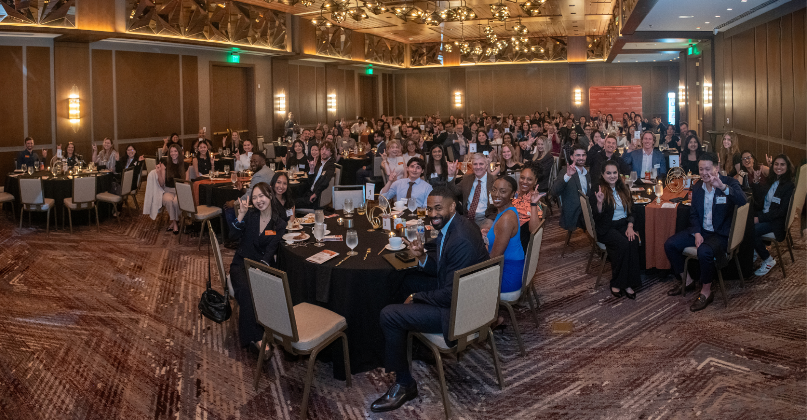 A large group shot of young professionals at a networking event, seated at round tables in a banquet room and smiling brightly to camera.