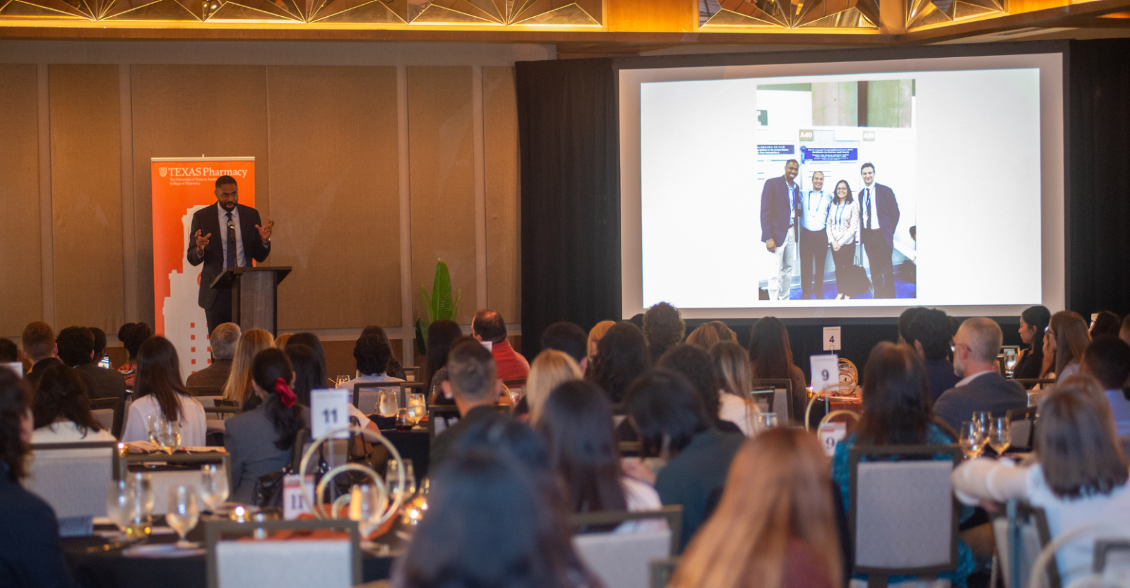 A professional man stands at the front of a room of seated attendees delivering a keynote speech. He speaks into a microphone at a podium beside a large presentation screen.