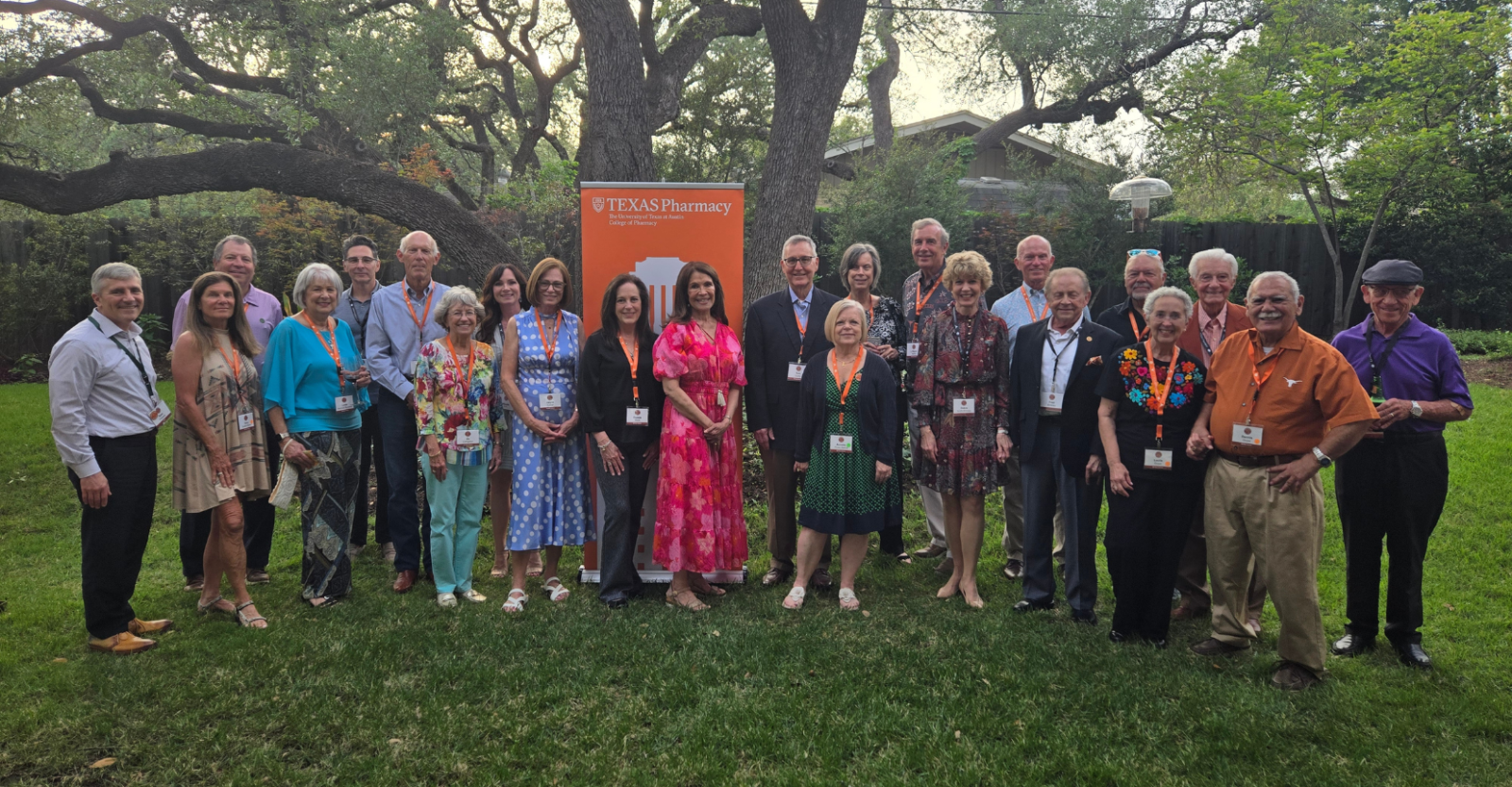 A group of people standing on grass in front of a "UT Texas Pharmacy" sign, under large trees. The group appears to be attending a formal or semi-formal event.