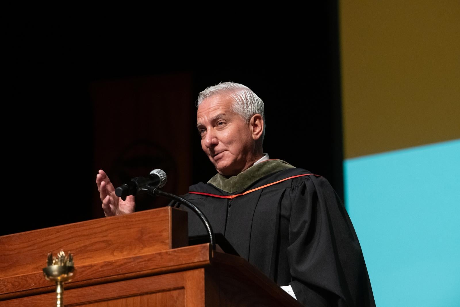 A man standing at a podium giving a speech in graduation robes.