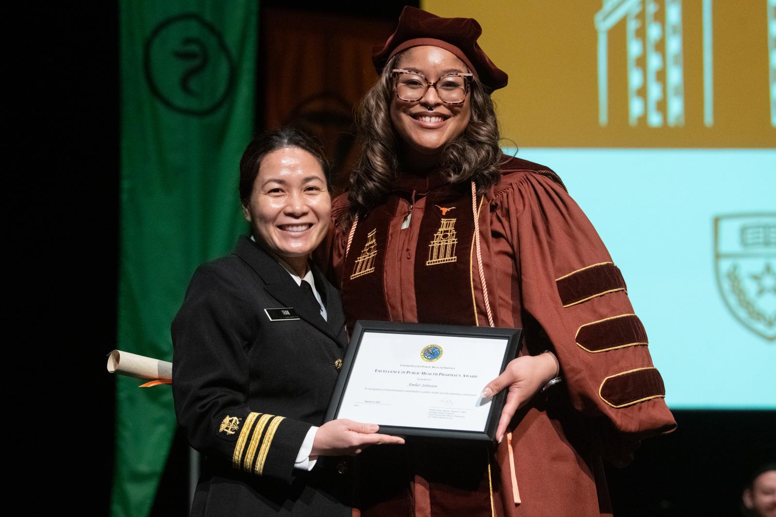 Two people standing on a graduation stage with an award certificate.