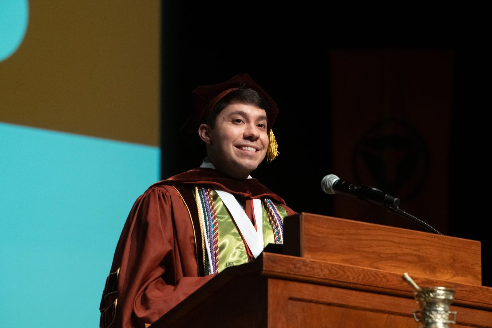 A man smiling while giving a speech in graduation robes.