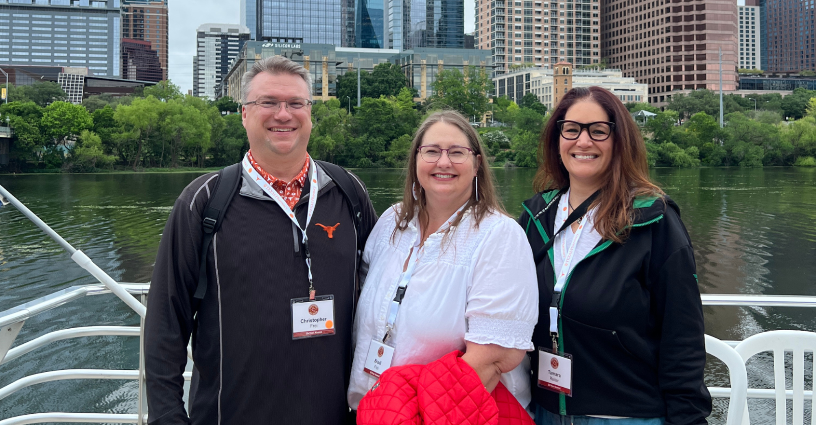 Three people are smiling on a boat with water and a cityscape in the background. They are wearing conference lanyards, and the person on the left has a Longhorn logo on their jacket. A red jacket is draped over the arm of the person in the middle.