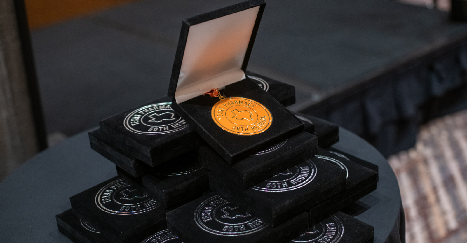 A stack of Texas Pharmacy 50-year gold medals lies on a table, with one award prominently displayed in an open box.