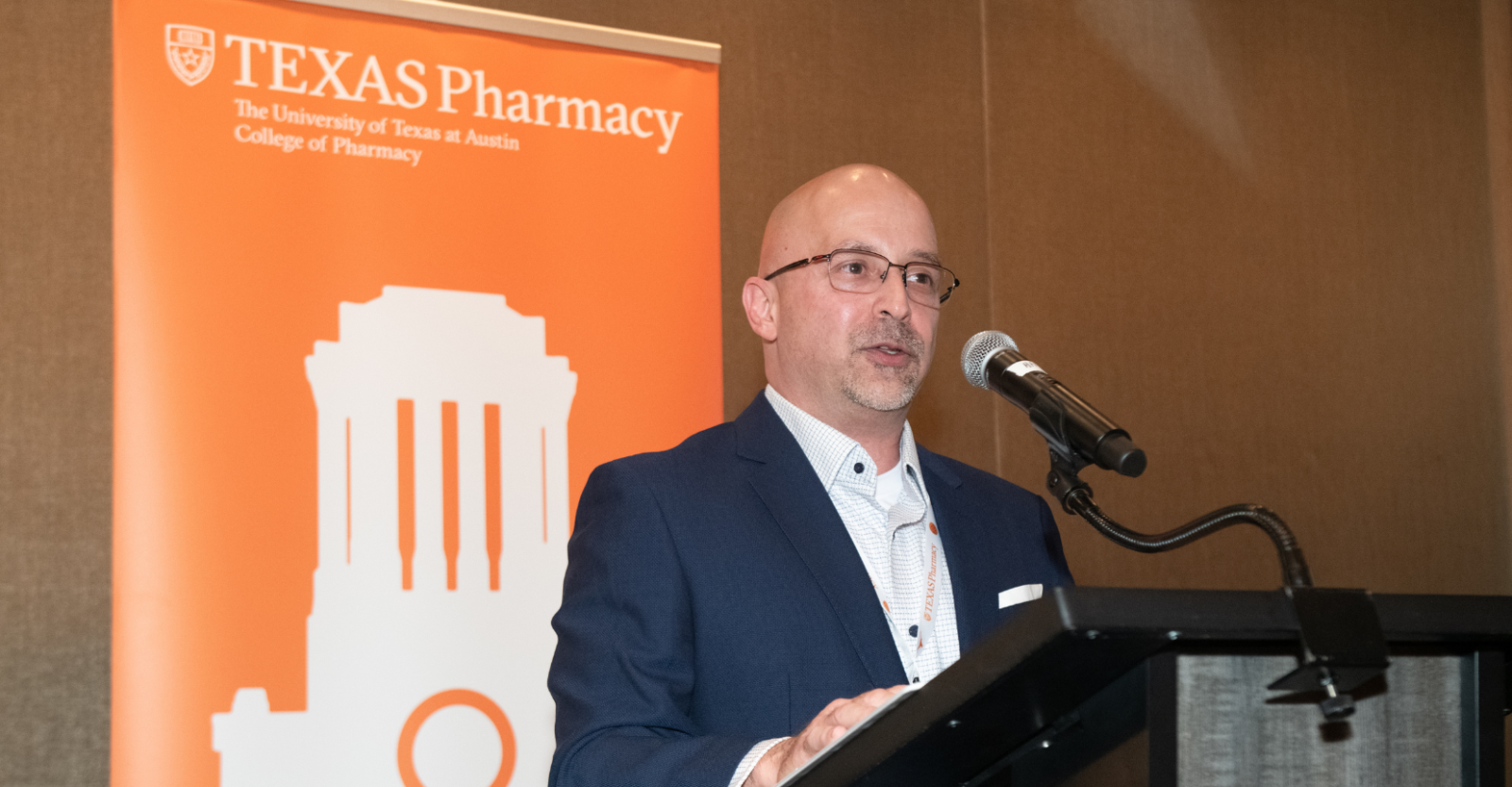 A person stands at a podium speaking into a microphone. Behind them is an orange banner with the Texas Pharmacy logo from the University of Texas at Austin College of Pharmacy.