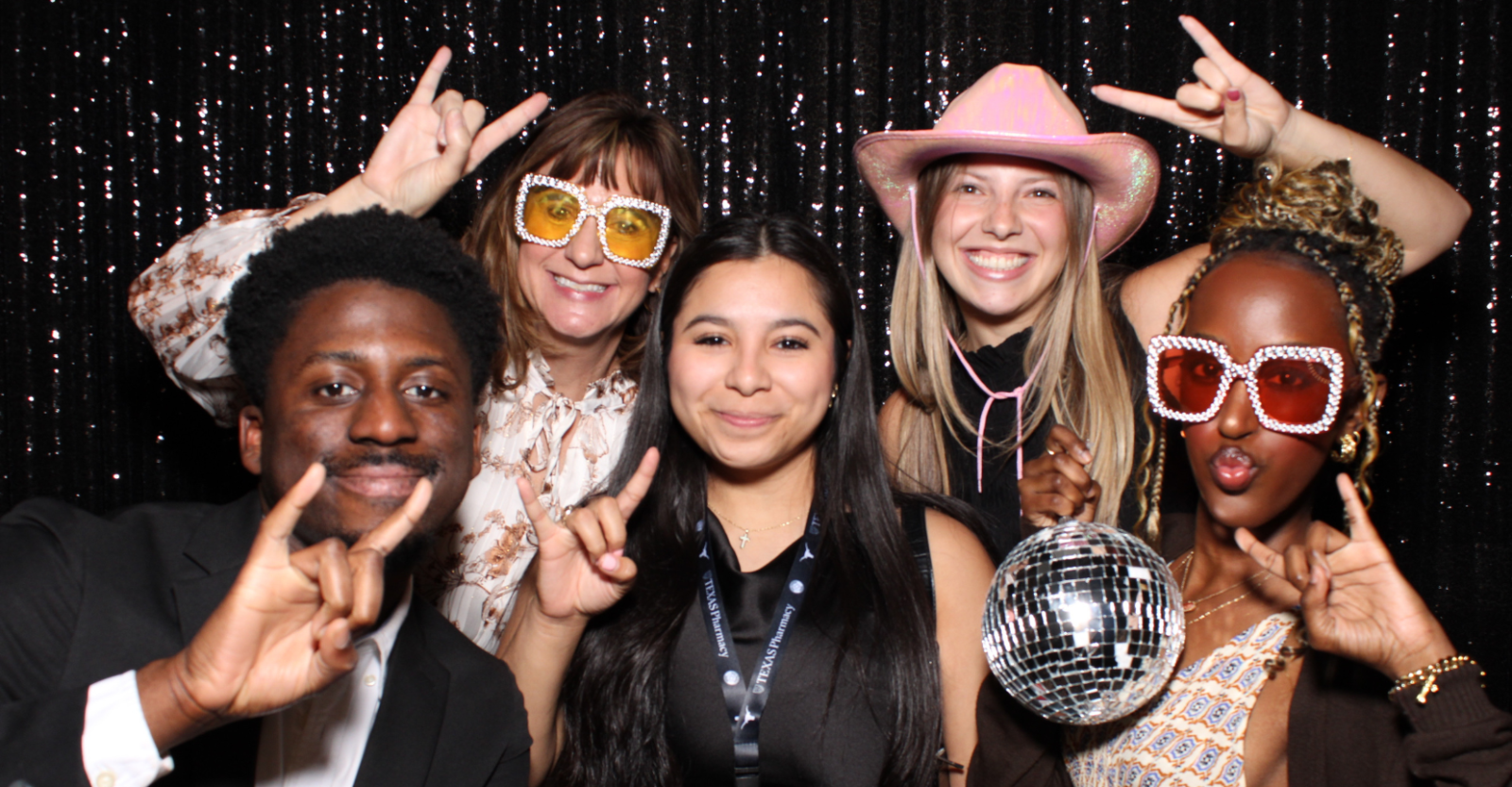 Five people posing cheerfully in front of a sequined backdrop, making hand signs. One person holds a disco ball, while others wear stylish sunglasses and hats. They appear to be having fun at a social event.