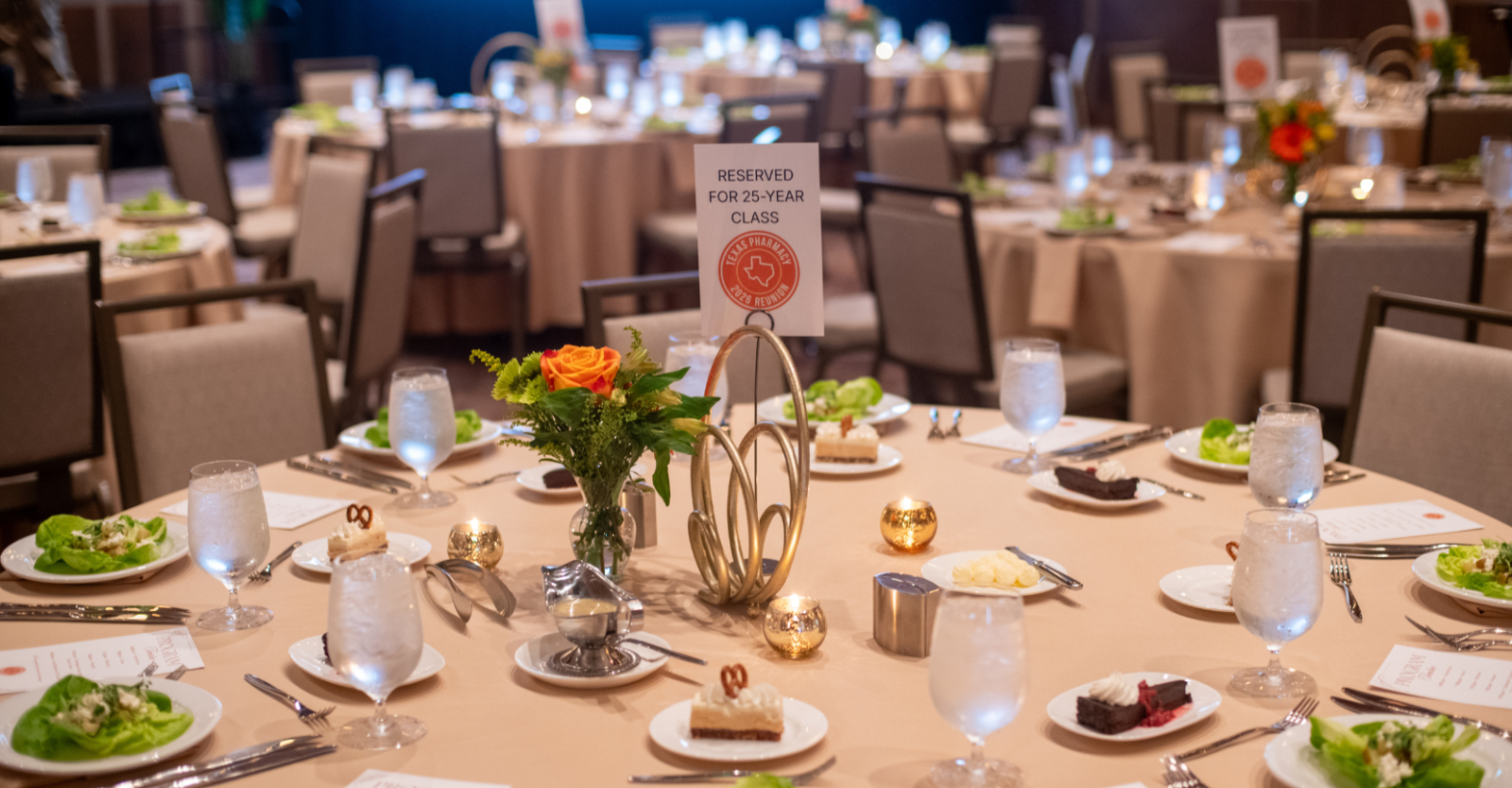 A formal dining setup in a large room, for a Class Reunion event. Multiple round tables are adorned with beige tablecloths and set with plates, silverware, and glasses of water. Each setting includes a salad, a piece of cake, and a small piece of chocolate. A centerpiece with a small floral arrangement decorates the middle of the table. A sign reads "Reserved for 25-Year Class" with the Texas Pharmacy logo. Other tables and chairs are visible in the background.