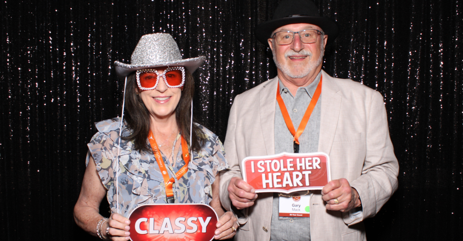 A couple stand together in a photo booth wearing cowboy hats and decorative glasses. They hold signs that read "CLASSY" and "I Stole Her Heart"