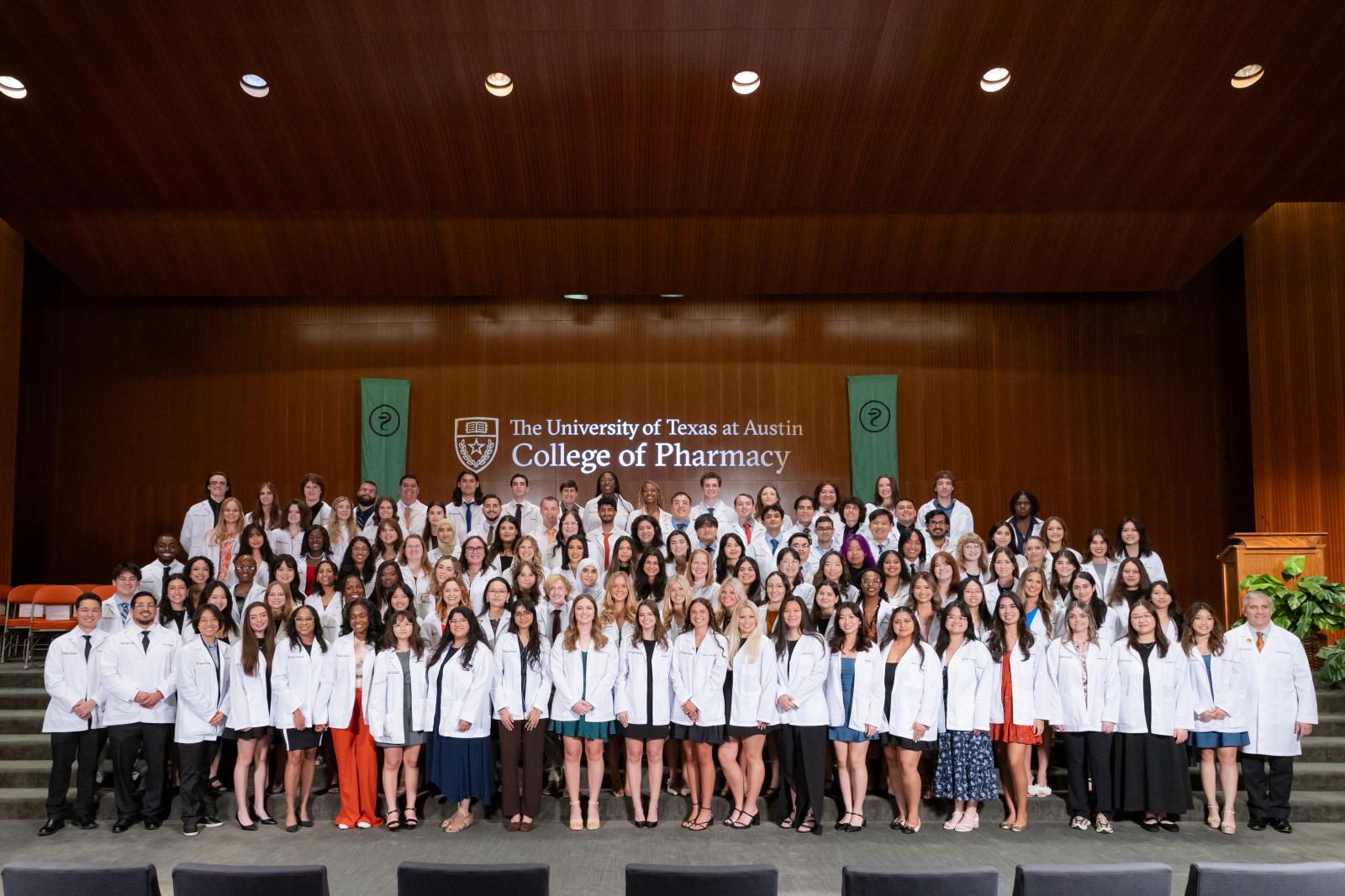 Group photo of a UT Austin College of Pharmacy class at the annual white coat ceremony. The students stand on stage, wearing their coats and smile brightly to camera.