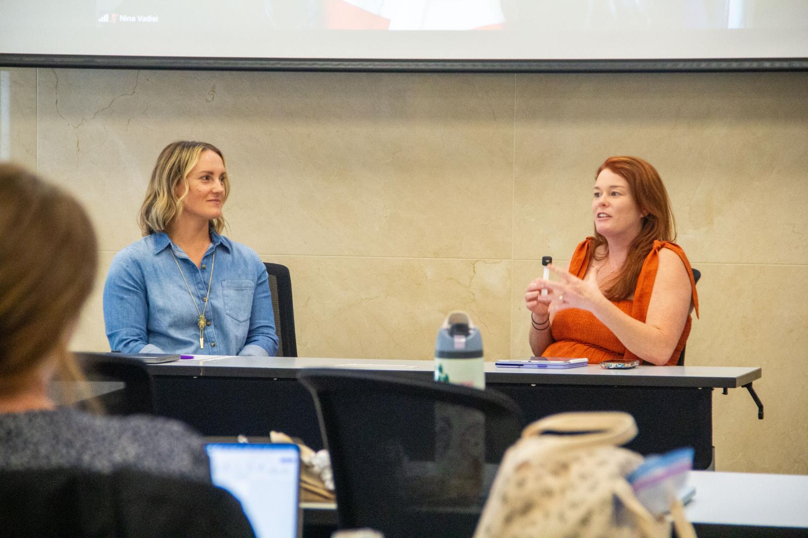 Two young female professors in casual dress sit together at a table and address a room of attendees.