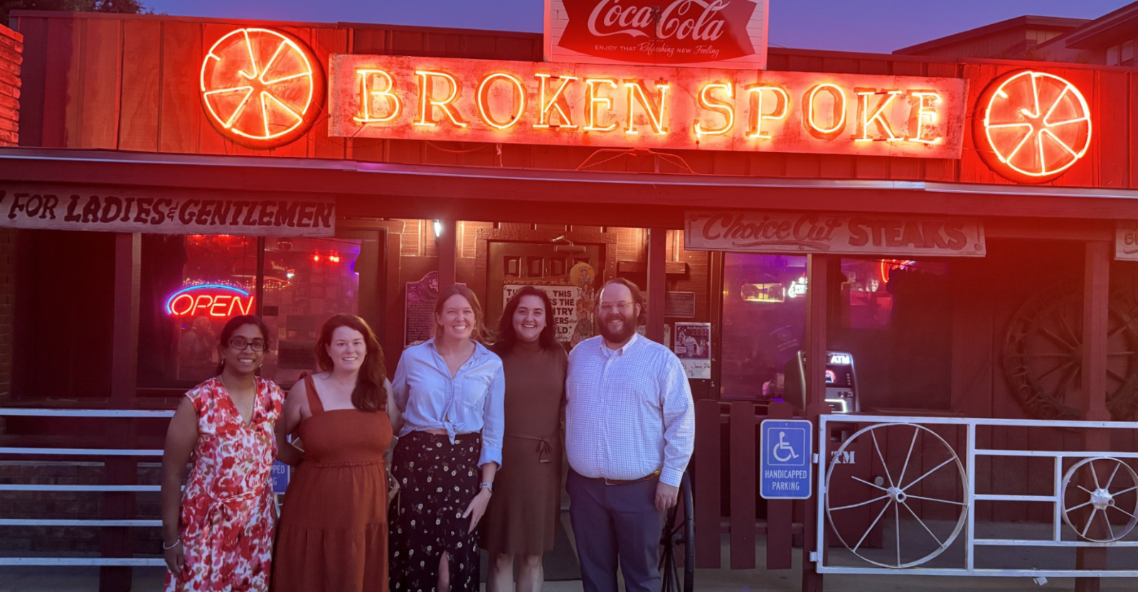 A group of five young professionals posing together in front of a neon bar sign that reads "Broken Spoke"