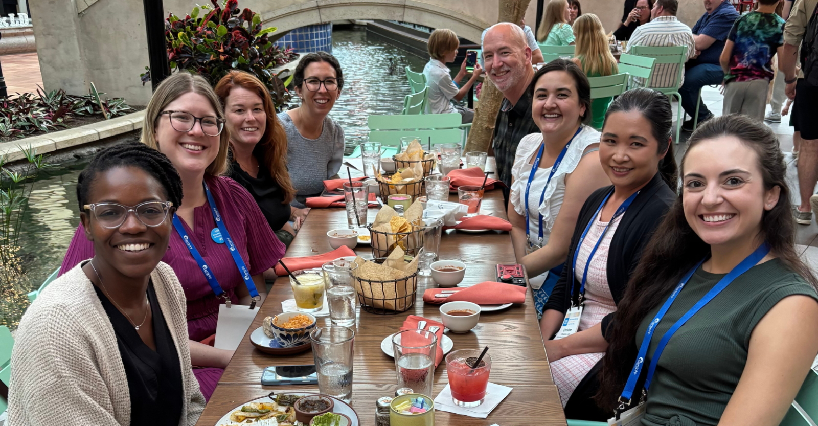 A large group of young professionals smile brightly to camera. They are sitting at a table at an outdoor restaurant, sharing a meal and drinks together.