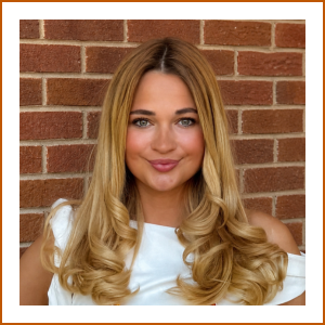 Headshot of student pharmacist Katherine. Katherine has curled, blonde hair and is wearing a white dress. She smiles softly to camera in front of a red brick wall.