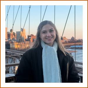 A headshot of student pharmacist Megan. She is smiling warmly to camera wearing. a black jacket and white scarf. She poses on a bridge with a cityscape at sunset in the background.