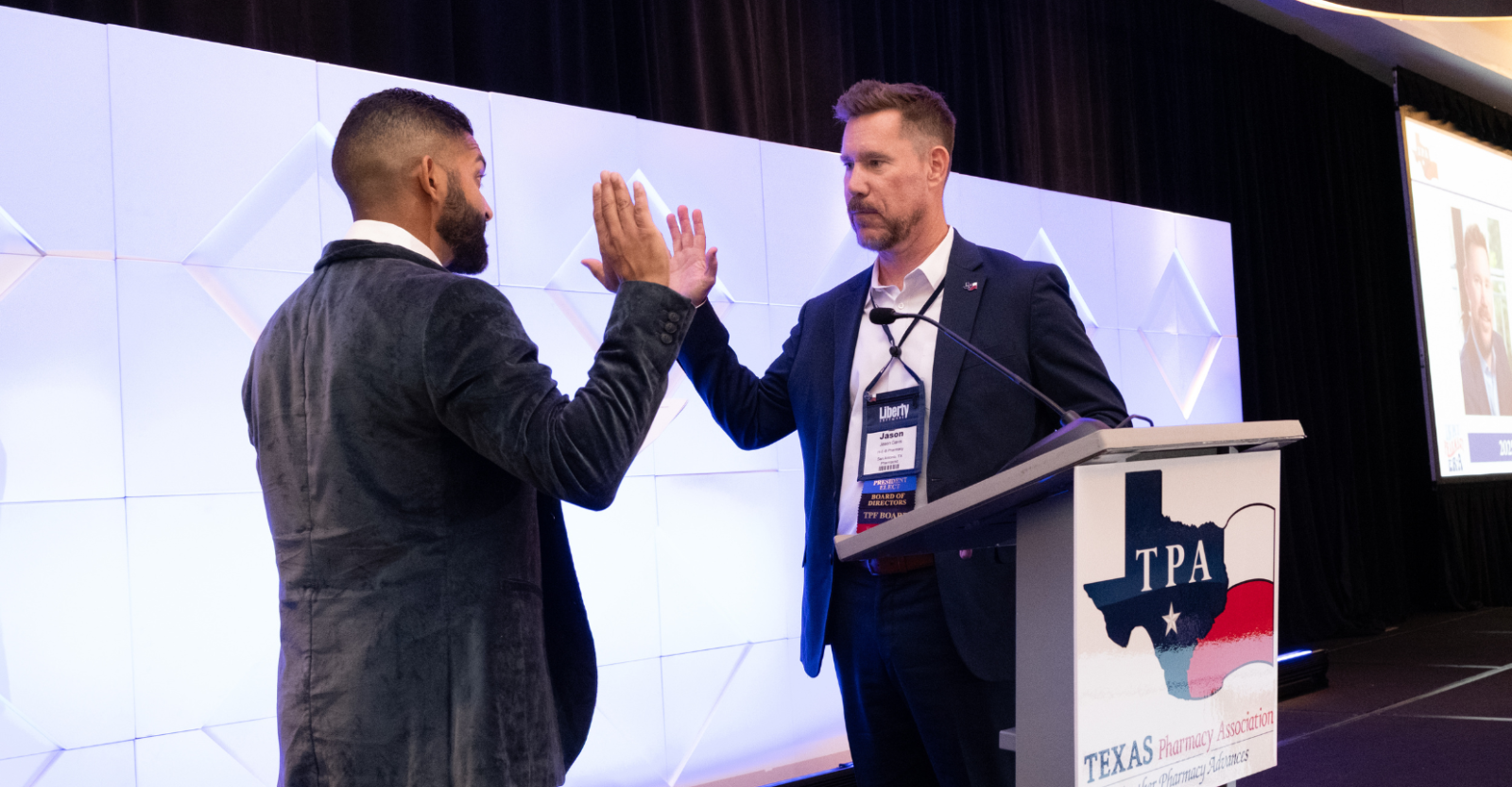 Two men in suits stand on stage during a swearing in ceremony at the Texas Pharmacy Association meeting.