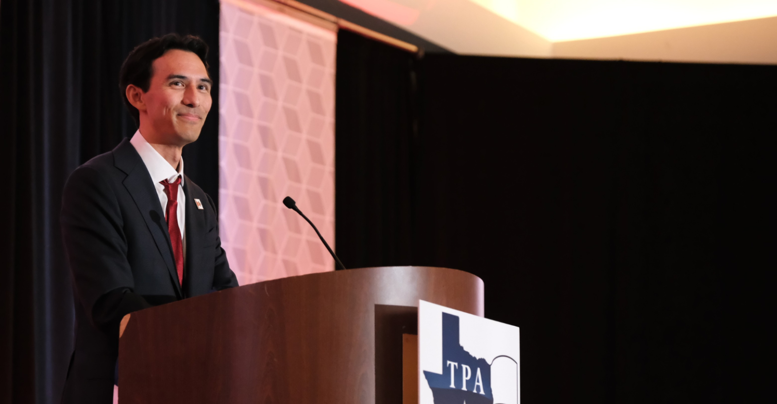 A man in a dark suit and red tie addresses an unseen audience on stage behind a Texas Pharmacy Association podium.