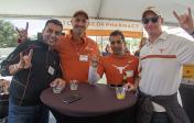 Four Pharmacy alumni smiling and giving hook em horns sign at cocktail table at Homecoming Tailgate event