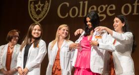 Student pharmacists standing on stage during the White Coat Ceremony. A female student with brown hair and a pink dress is receiving her white coat from another woman while colleagues look on, smiling.