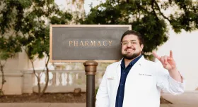 A headshot of student pharmacist Erik Coronado. Erik poses outside the Pharmacy building by the Pharmacy sign. He has brown hair and a brown beard and glasses. Erik holds his hands with "horns up" and smiles to camera in his white coat.