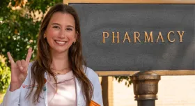 A woman standing in front of a pharmacy sign and giving the Hook 'em Horns hand gesture.