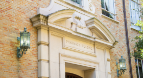 Front facade of the College of Pharmacy building at UT Austin. Multi-colored brick surrounds a decorated stone archway above a wooden door.