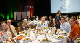 A group photo of Texas Pharmacy alumni, faculty and friends sitting together at a round gala table during a college event. All are smiling brightly to camera.