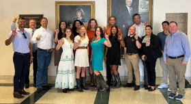 A group of staff and faculty from the UT Austin College of Pharmacy San Antonio team pose together in front of painted portraits in a hallway.