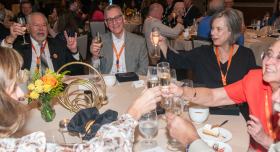A group of people seated around a table at a formal event, raising glasses in a toast. The table has floral centerpieces and place settings. Several attendees are wearing lanyards.