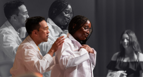 On stage at a white coat ceremony, a pharmacy student puts on her first white coat with the help of a faculty member.