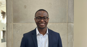 A headshot of graduate student Ayobami standing infront of a brick wall, wearing a suit and a conference lanyard. He wears glasses and smiles brightly to camera.