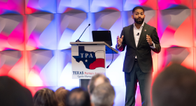 A person is presenting on stage at the Texas Pharmacy Association event. They stand beside a podium displaying the TPA logo and a map of Texas. The background has a geometric design lit with red and blue lights. An audience is seated in the foreground.