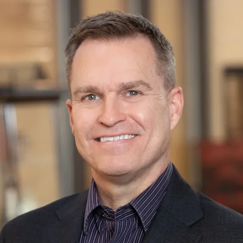 John Reineke wearing a suit coat and collared shirt and smiling.