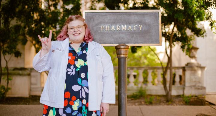 Student pharmacist Hannah McCullough standing with the Pharmacy building sign in white coat.
